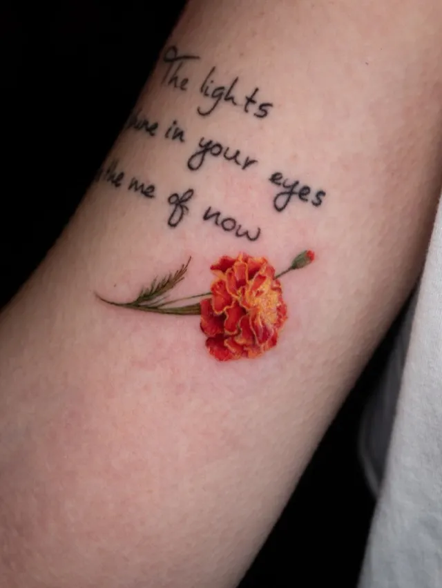 Farbiges Tattoo: leuchtend orange-rote Ringelblume mit feinem Schriftzug auf dem Oberarm