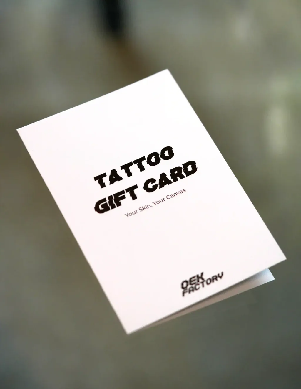 Gefaltete Geschenkkarte der OEK Factory in Weiß mit dem Aufdruck Tattoo Gift Card, Your Skin Your Canvas