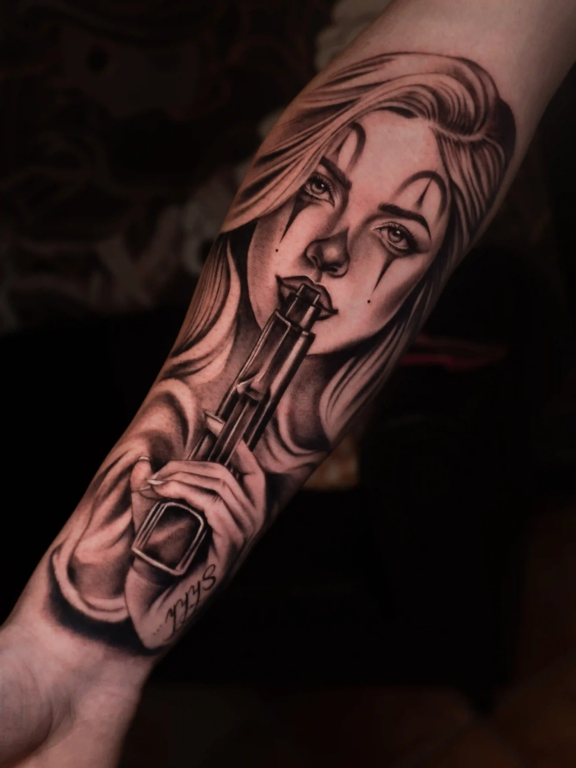Black and grey realism chicano style tattoo of a female portrait with clown makeup holding a gun to her lips, executed with dramatic lighting and smooth shading on the forearm.