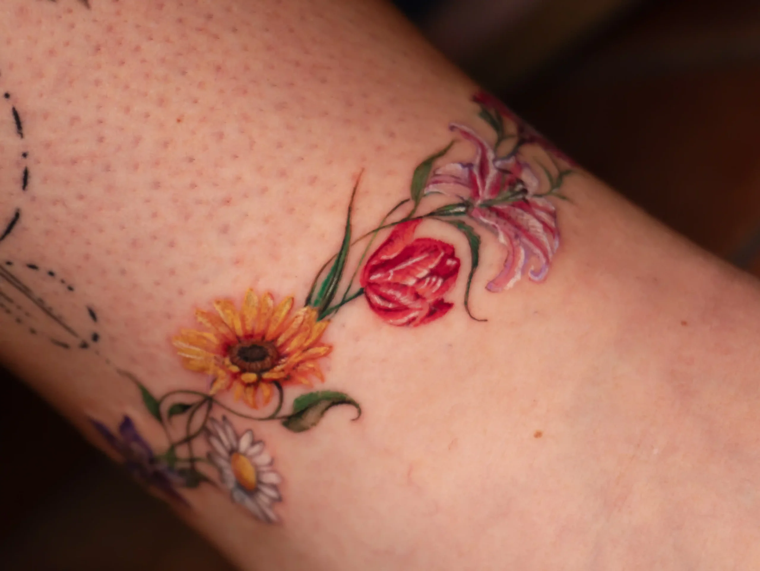 Colorful microrealism floral tattoo featuring a mix of daisies, tulips, lilies, and sunflowers in a fine-line watercolor style on the ankle.
