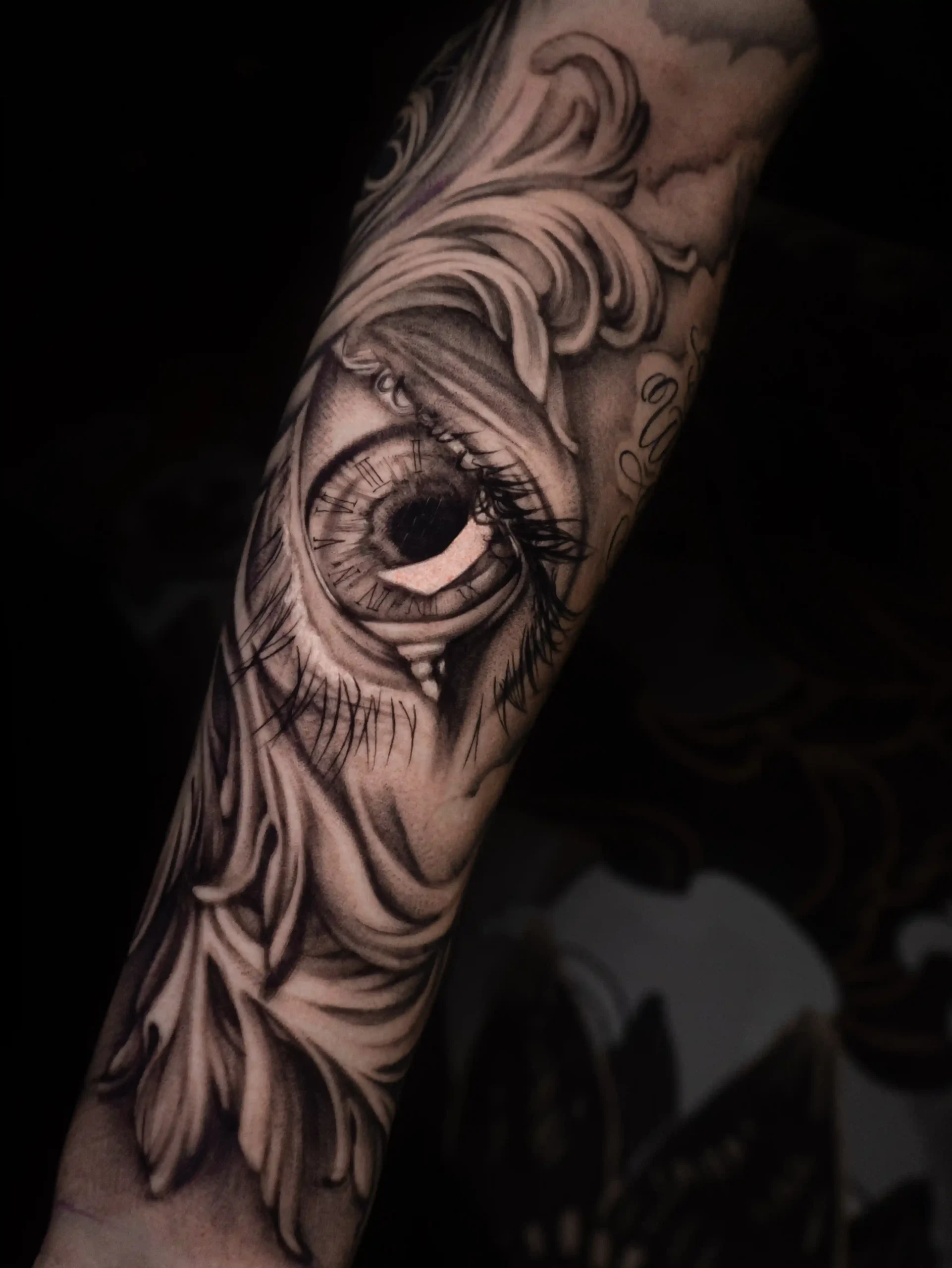 Realistic black and grey tattoo of a detailed human eye surrounded by ornamental baroque-style flourishes, emphasizing depth and contrast on the forearm.